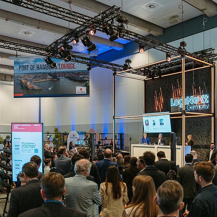 Besucherinnen und Besucher verfolgen eine Präsentation auf einer Logistik-Fachveranstaltung vor einer Bühnen- und Ausstellungsfläche.
