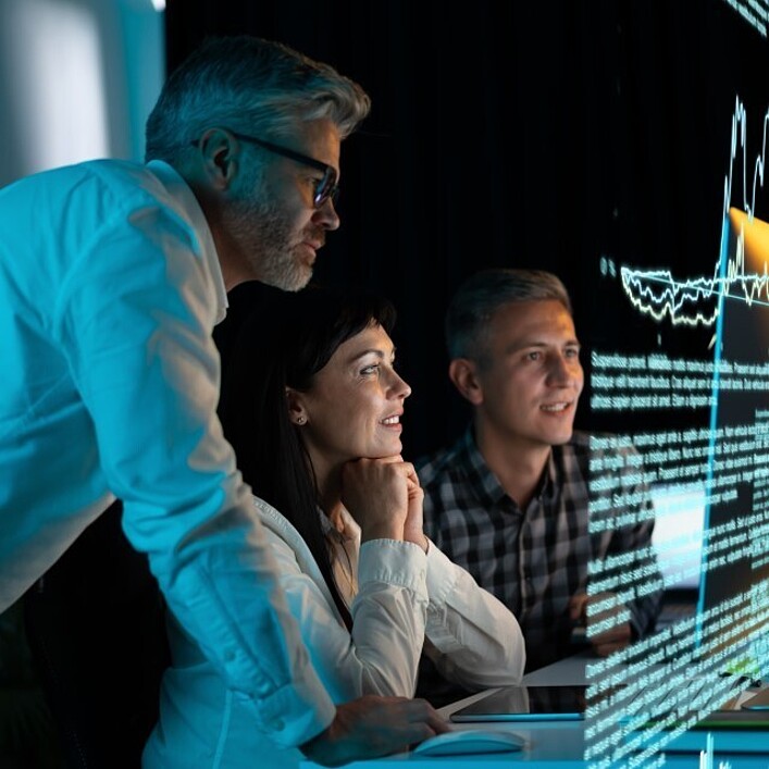 Mehrere Personen sitzen vor großen digitalen Analyse-Displays mit grafischen Daten und Texten, während eine weitere Person sich über den Tisch lehnt und auf den Bildschirm zeigt.