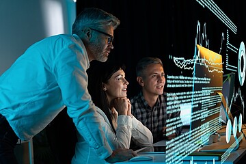 Mehrere Personen sitzen vor großen digitalen Analyse-Displays mit grafischen Daten und Texten, während eine weitere Person sich über den Tisch lehnt und auf den Bildschirm zeigt.